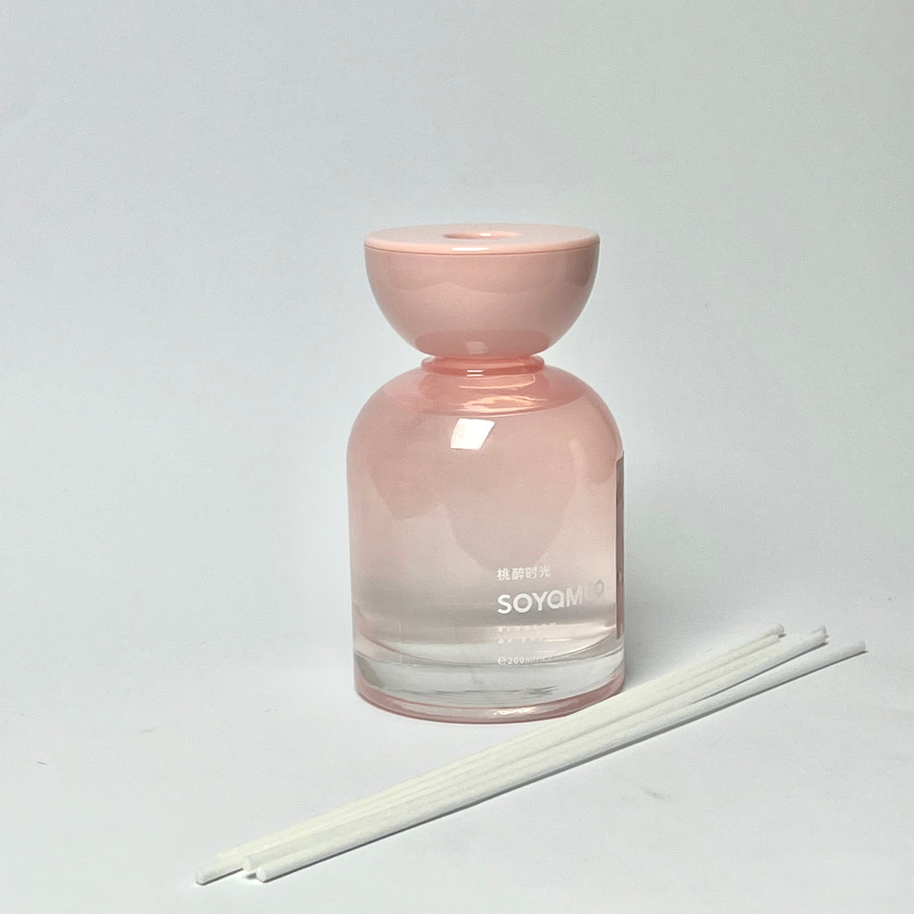 A pink glass reed diffuser bottle with a matching pink stopper, shown on a light gray background with three white reeds in front.