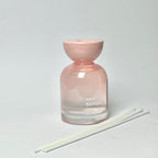 A pink glass reed diffuser bottle with a matching pink stopper, shown on a light gray background with three white reeds in front.