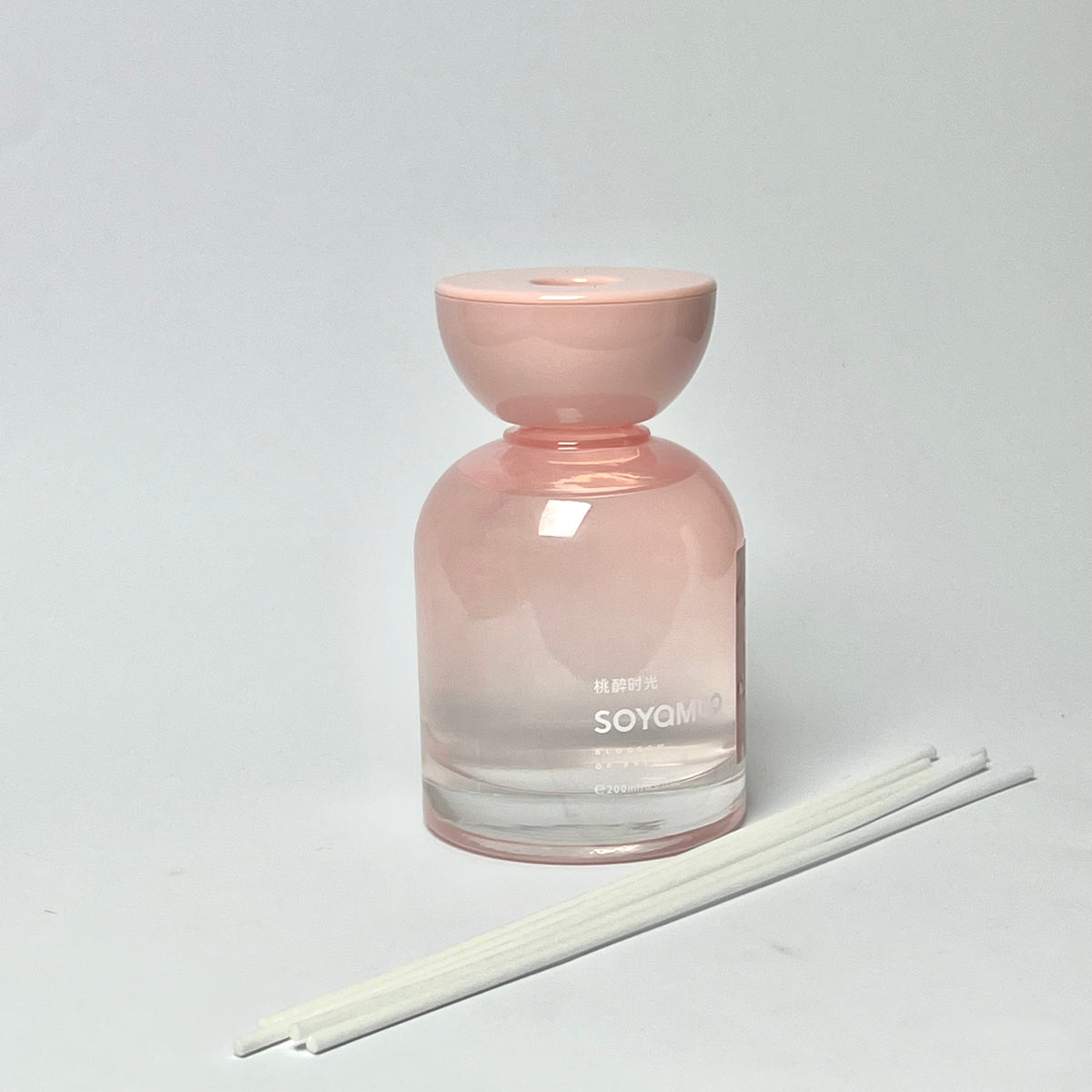A pink glass reed diffuser bottle with a matching pink stopper, shown on a light gray background with three white reeds in front.