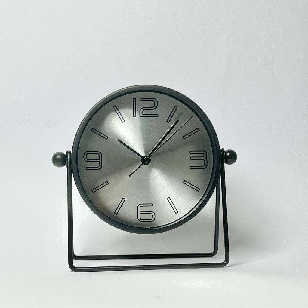 A modern round desk clock with a brushed silver face and black frame, supported by a black metal stand against a plain white background.