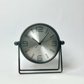 A modern round desk clock with a brushed silver face and black frame, supported by a black metal stand against a plain white background.
