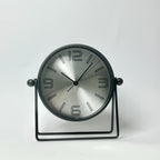 A modern round desk clock with a brushed silver face and black frame, supported by a black metal stand against a plain white background.