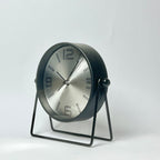 A modern cylindrical desk clock with a brushed silver face and black casing, supported by a minimalist black metal stand against a light gray background.