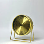 A modern round table clock with a brushed gold finish, featuring black Arabic numerals and hands on a minimalist wire metal stand, set against a plain light gray background.