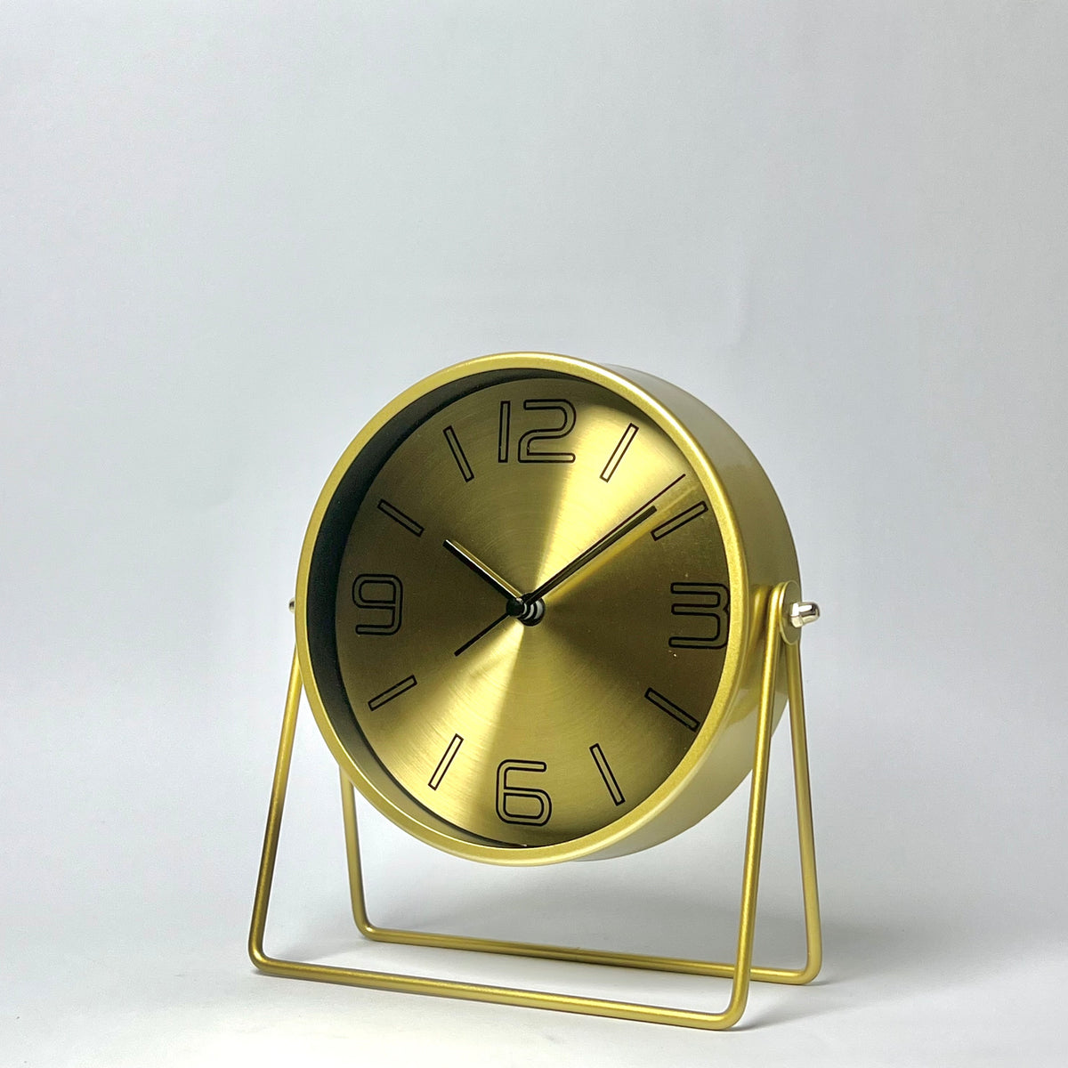 A modern round table clock with a brushed gold finish, featuring black Arabic numerals and hands on a minimalist wire metal stand, set against a plain light gray background.