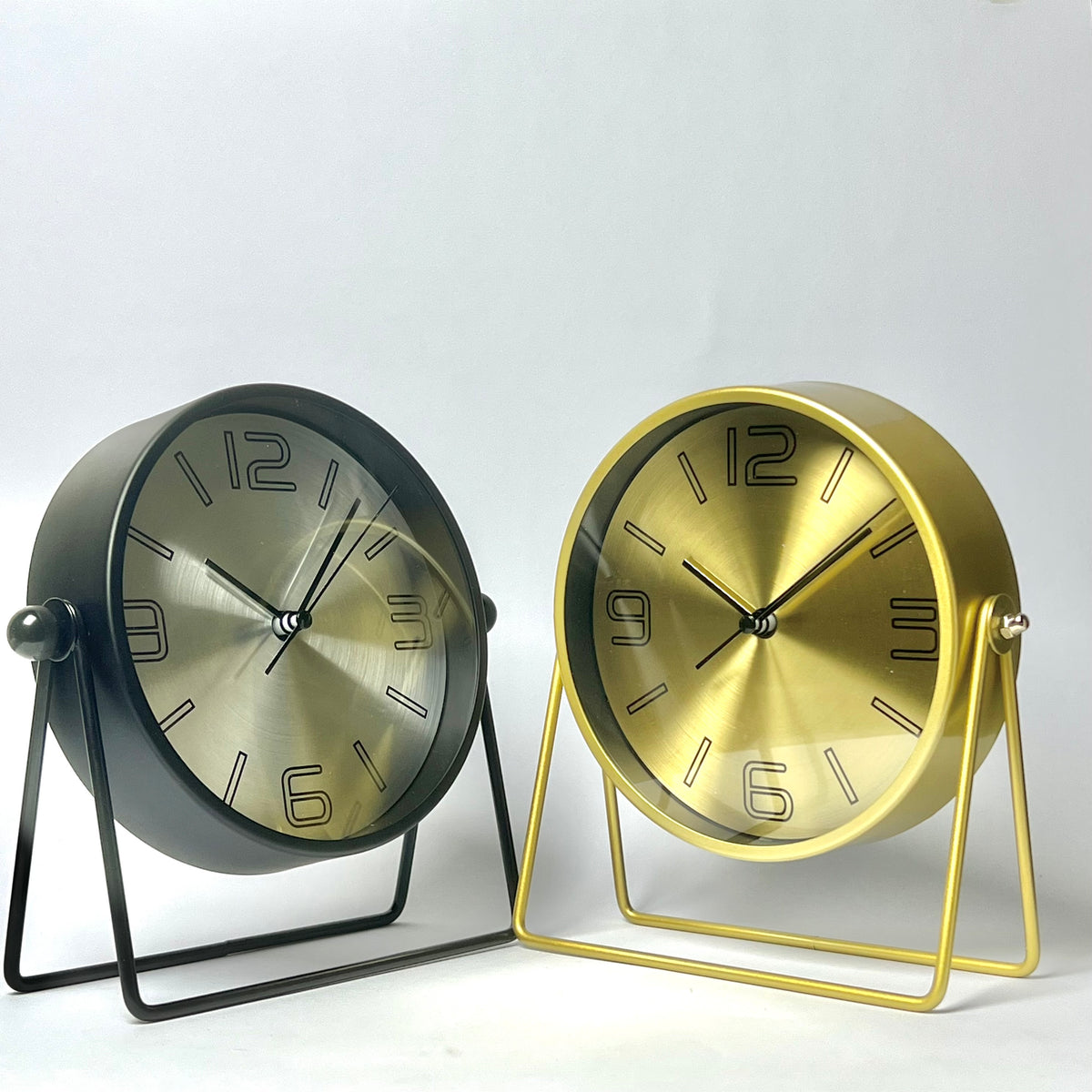 Two round tabletop clocks with brushed metal faces and black numerals, shown side by side on simple metal stands against a light gray studio background. The left clock has a dark gray body and stand, and the right clock has a gold body and stand.