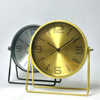 Two round tabletop clocks with minimalist wire stands on a light gray background. The front clock has a brushed gold dial and gold metal frame, while the rear clock has a brushed silver dial and black metal frame.