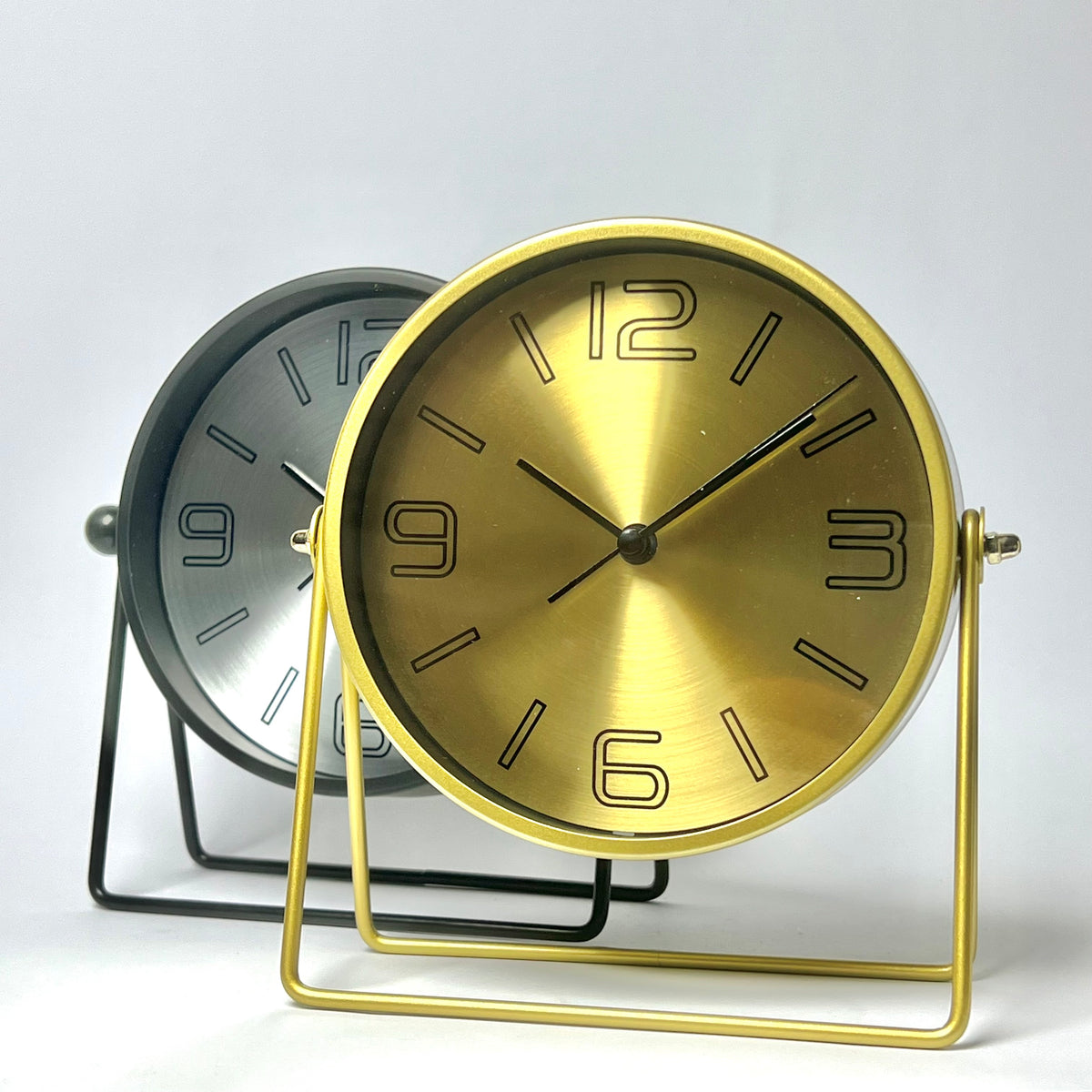 Two round tabletop clocks with minimalist wire stands on a light gray background. The front clock has a brushed gold dial and gold metal frame, while the rear clock has a brushed silver dial and black metal frame.