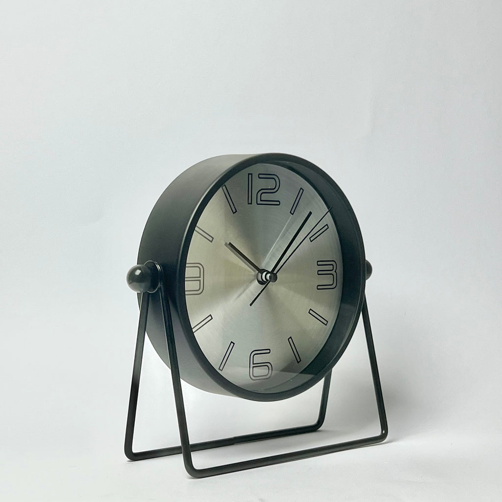 A modern round desk clock with a brushed silver dial and black frame, supported by a black metal U-shaped stand against a plain white background.