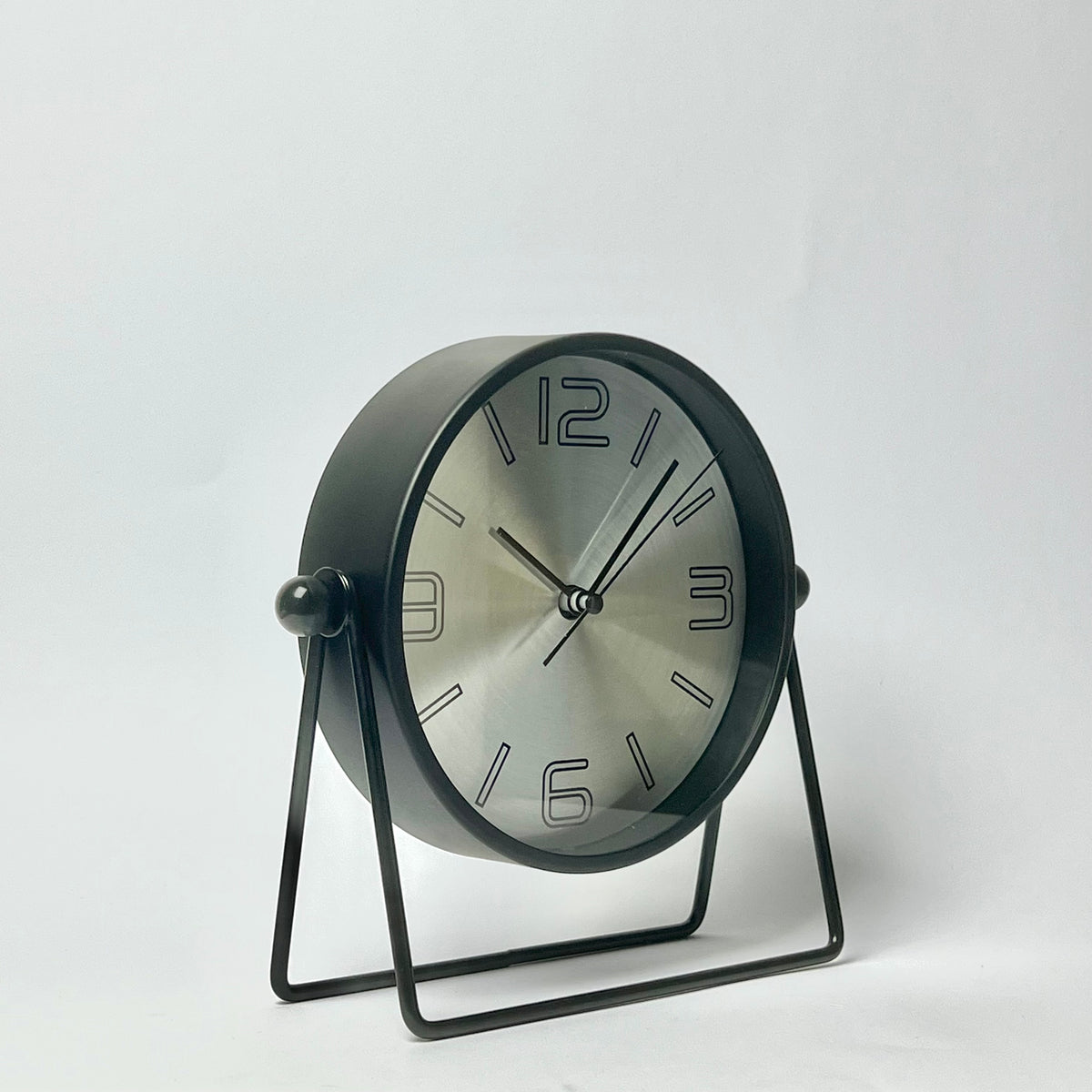 A modern round desk clock with a brushed silver dial and black frame, supported by a black metal U-shaped stand against a plain white background.