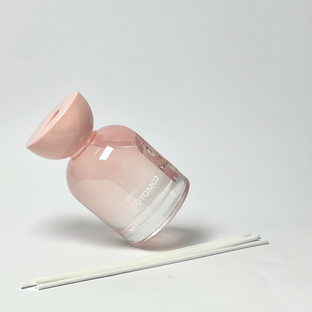 A pink glass reed diffuser bottle with a matching pink stopper, shown on a light gray background with three white reeds in front.